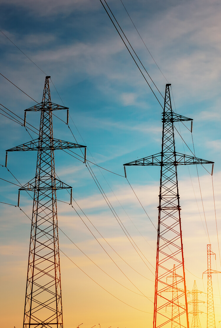 Two electricity transmission towers against a sky transitioning from blue to orange, symbolizing power and electrical systems.